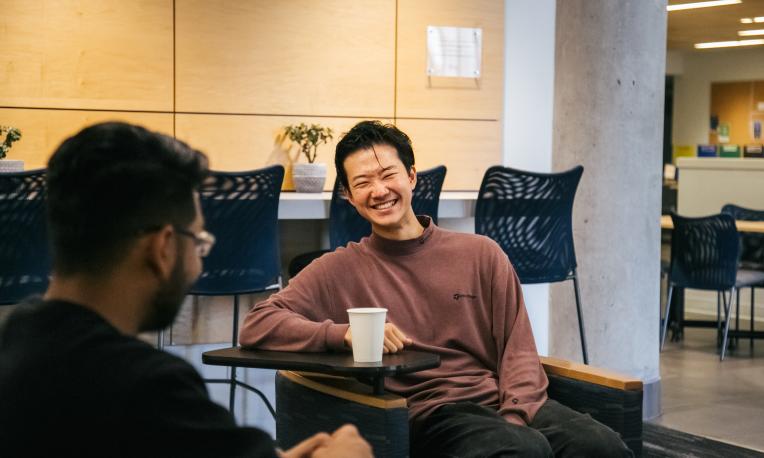 Two males sitting, talking, and smiling over coffee.