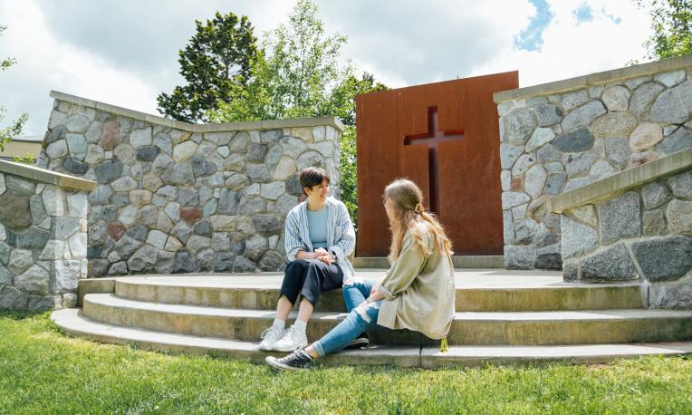 Two female students sitting on the steps of the Langley campus outdoor chapel talking. There is a cross in the background.