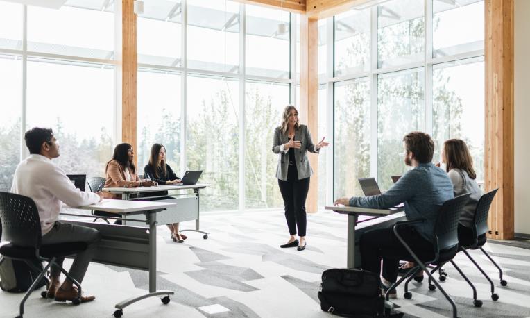 Teacher speaking to students sitting in a classroom with windows