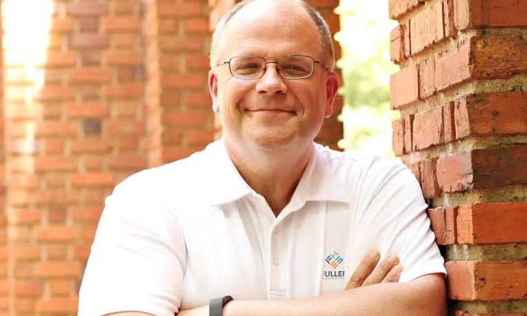 person leaning on brick wall with arms crossed, smiling at the camera