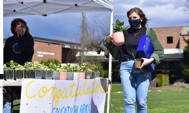 A student in a mask holds a potted plant and walks past a booth of plants.