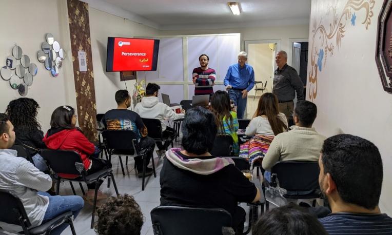 Audience listening to speakers in a small room with a presentation screen.