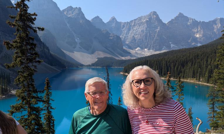 Two smiling people stand by a turquoise lake with mountains and trees in the background.