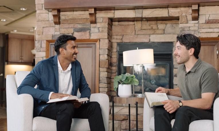 Two men chatting and smiling in a cozy living room with a fireplace.