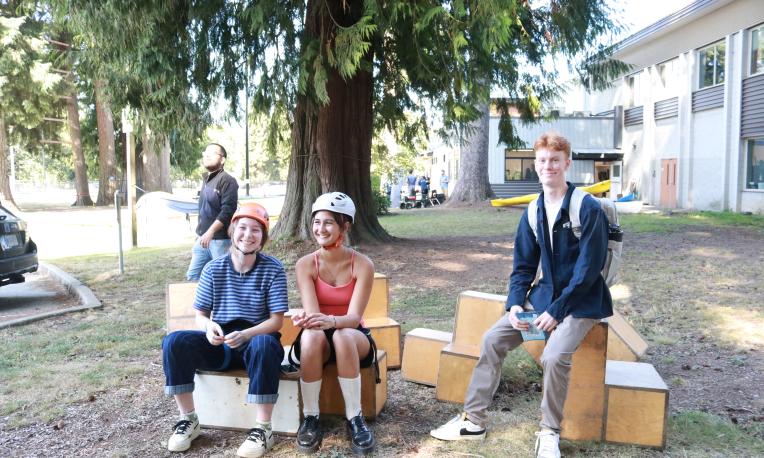 Three people sitting on wooden benches under a tree, two wearing helmets, one in a suit.