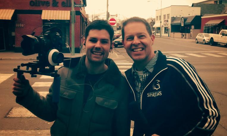 Two smiling men on a street, one holding a camera rig.