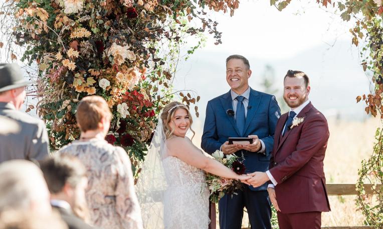 Mike and bride and groom smiling at an outdoor wedding ceremony.