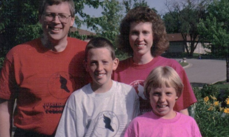Smiling family of four standing outdoors, wearing casual shirts.