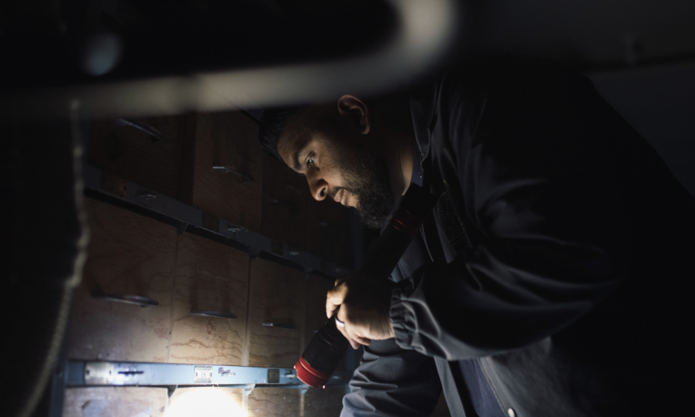 Man inspecting a dark area with a flashlight, wearing a jacket.