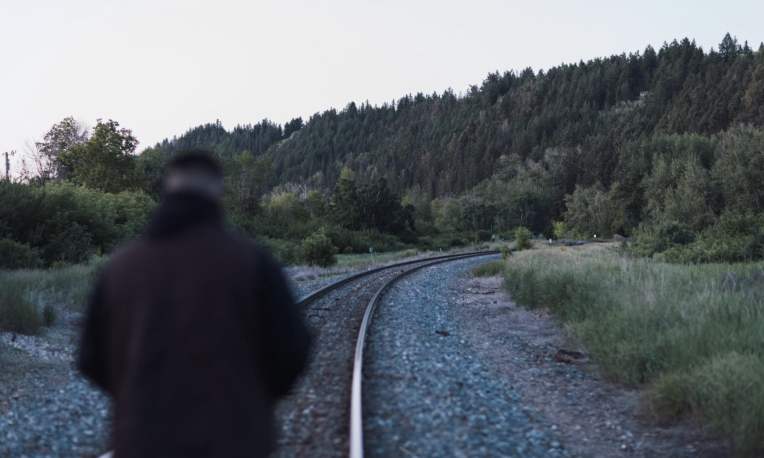 A person walks along a curved railroad track by a forest.