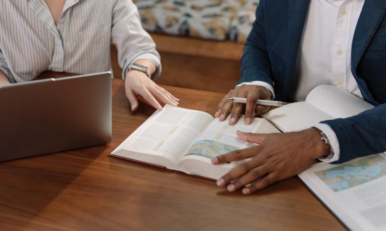 Two people studying a Bible and using a laptop at a wooden table.