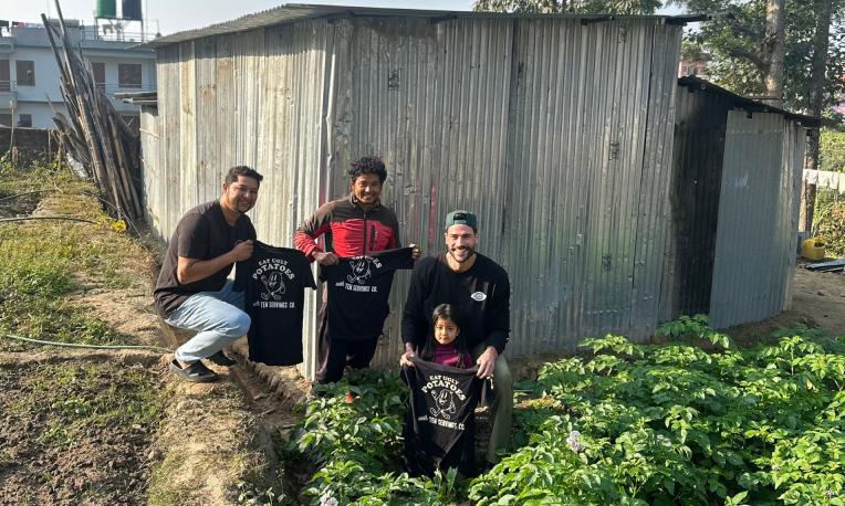 Four people holding black shirts in a garden by a metal shed, sunny day.