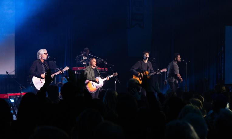 Band performing on stage with guitars, blue lighting, and an audience.