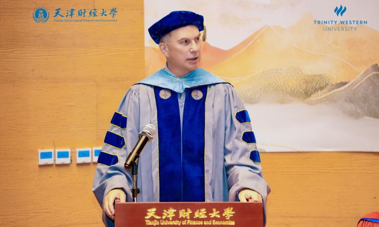 Man in academic regalia speaks at podium with university logos behind.