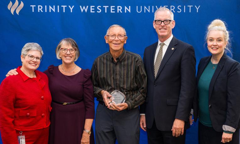 Five people stand smiling, one holding an award, against a blue backdrop labeled Trinity Western University.