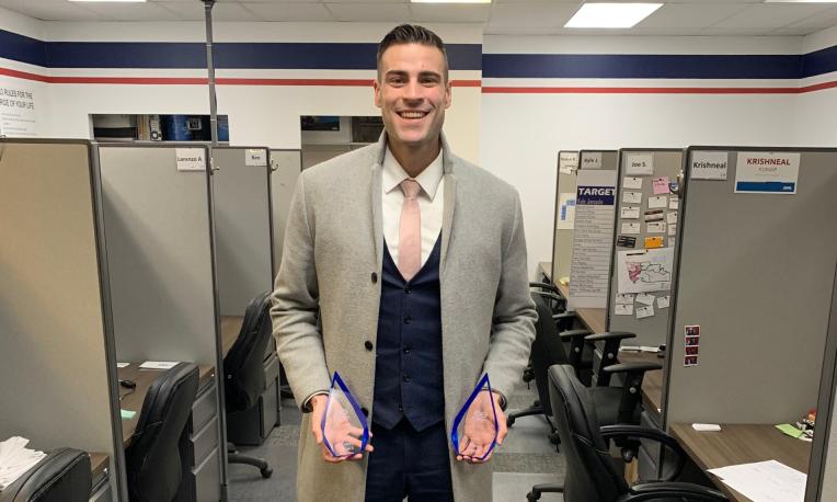 Smiling man in an office with cubicles, holding two blue awards.
