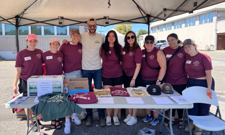 Group of people in maroon shirts under a canopy at an outdoor table with various items.