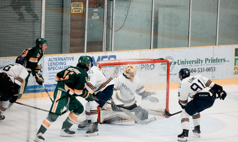 Hockey players battle near the goal as the goalie in white attempts to block a shot from a player in green. Two other players in white and one in green are close to the net during the play.
