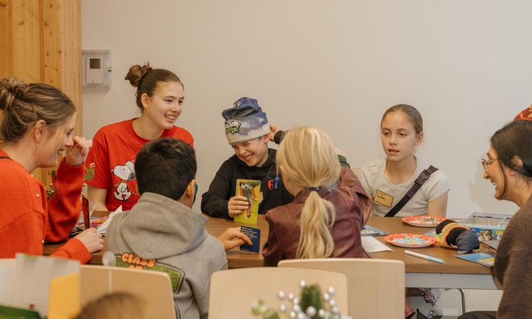 Young children and adults at a table, laughing and playing Christmas games.