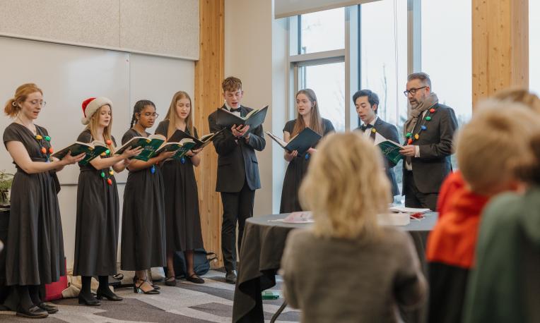 TWU Chamber Choir singing with songbooks in hands and colourful plastic Christmas lights draped around their shoulders.