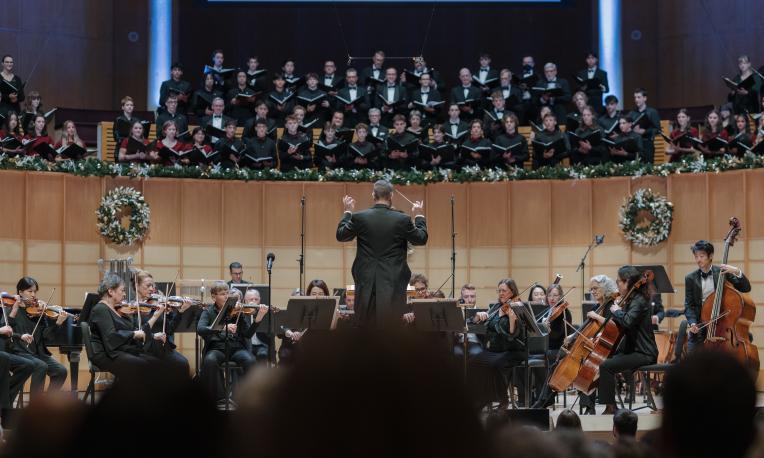 A conductor leads an orchestra on stage during a Christmas concert, with musicians playing string instruments in the foreground. Behind them, a large choir stands on an elevated platform, and the stage is decorated with festive wreaths and garlands.