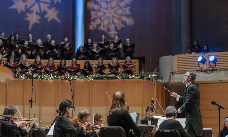A conductor leads an orchestra during a holiday concert in a large auditorium. The orchestra is seated in the foreground with string musicians playing, while a choir stands on an elevated platform in the background, holding sheet music. The stage is decorated with garlands and festive lights, and large snowflake patterns are projected onto the wall behind the choir, creating a seasonal atmosphere.