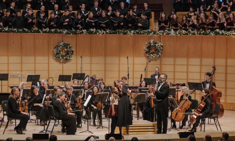 A soloist is positioned near the conductor, and behind them, a large choir is arranged on tiered seating. The stage is decorated with festive wreaths and garlands, creating a warm seasonal atmosphere.