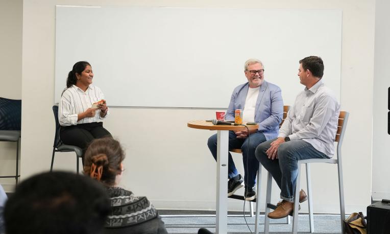 Three people sit at the front of a room in a panel-style discussion, with two seated at a tall round table and one seated to the side. An audience listens from the foreground, and a large whiteboard is visible behind the speakers.