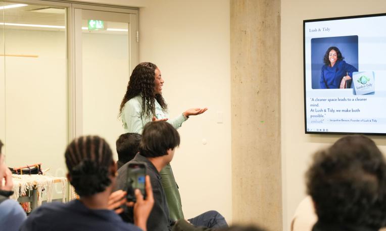 A Black woman stands at the front of a room giving a presentation to an audience, gesturing toward a screen displaying a slide about a cleaning business. Several attendees sit facing the presenter, some taking photos or notes.
