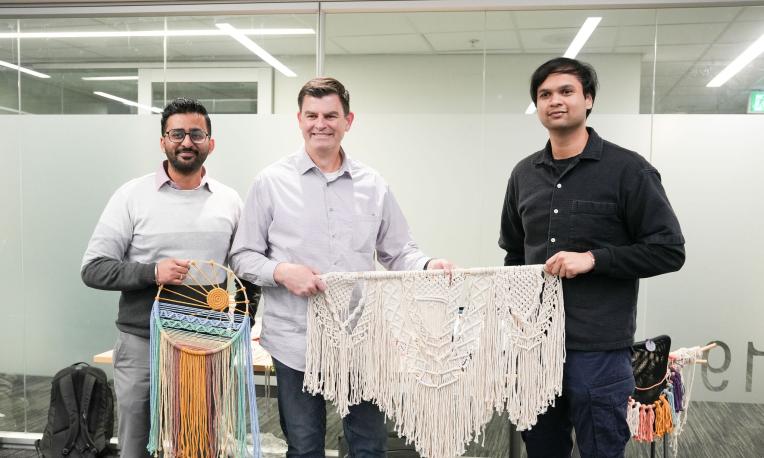 Three people stand indoors holding handmade woven artworks, including a colourful dreamcatcher-style piece and a large macramé wall hanging. 