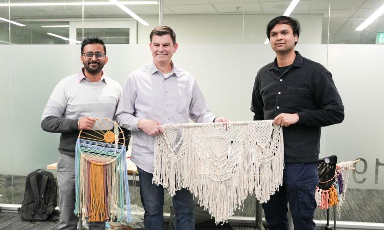 Three people stand indoors holding handmade woven artworks, including a colourful dreamcatcher-style piece and a large macramé wall hanging. 