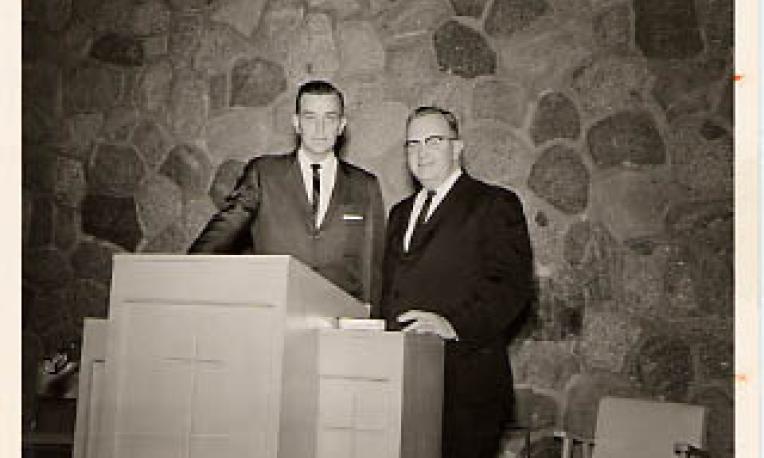 Photograph of founding president Calvin Hanson posing behind the pulpit on stage in the chapel with Harry Evans President of Trinity College and Trinity Evangelical Divinity School and guest speaker at the TJC Library dedication