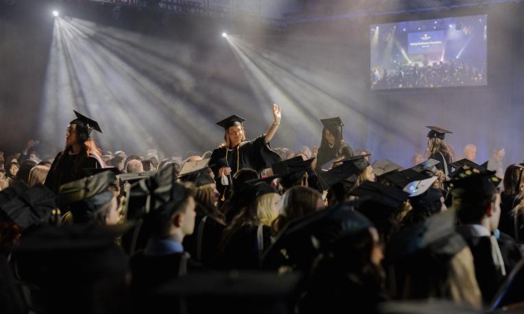 graduate walking in a cap and gown in a room with spotlights and the audience around her