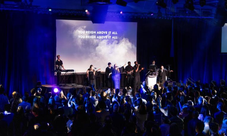 people singing on stage facing a crowd with a large screen behind them