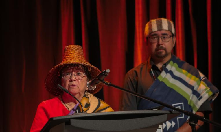 Patti Victor Switametelót speaking at a podium with a man standing behind her