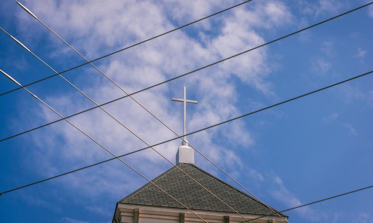 Cross at the top of a steeple in front of a blue sky with clouds and electrical wires in front