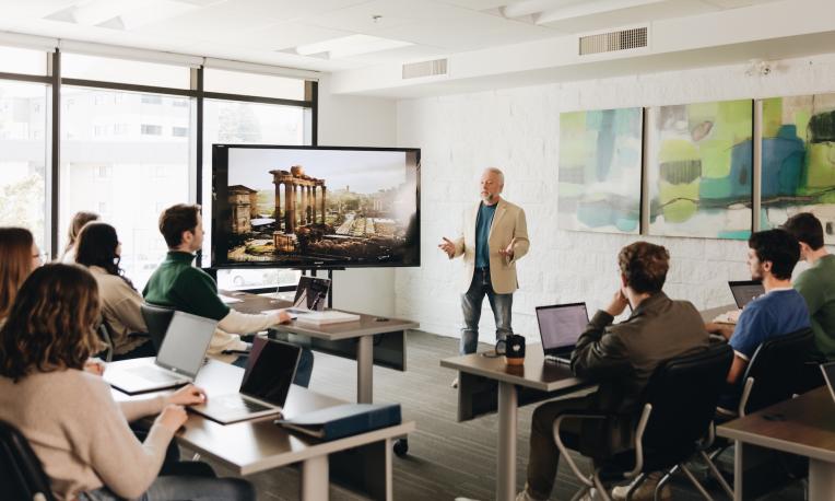 Instructor presenting to a classroom of students seated at tables with laptops in a bright TWU learning space. A large display screen shows an image of ancient ruins while students participate in the lecture.