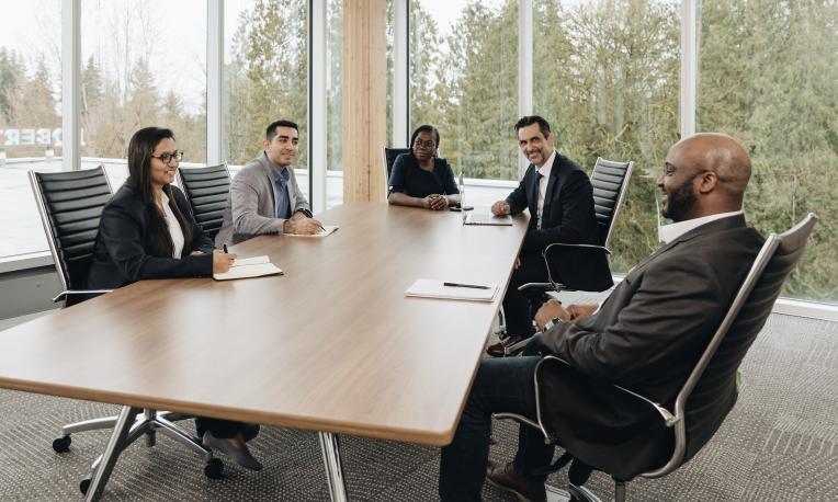 Five adult learners meet around a conference table in a bright, windowed room.