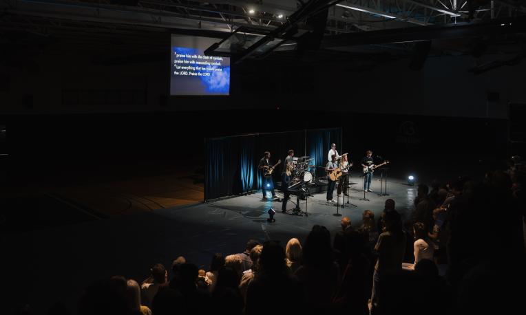 A worship band performs on stage in a dimly lit gym while a crowd gathers during TWU Homecoming.