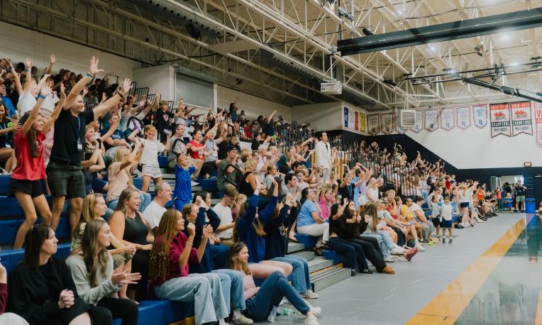 A large crowd of spectators fills the bleachers inside a university gymnasium, with many people raising their hands and cheering during a campus event. Championship banners hang on the wall above the court, and bright overhead lights illuminate the packed stands.