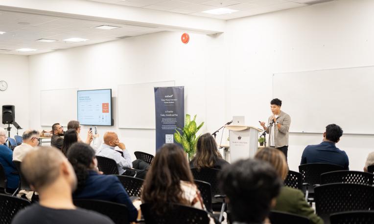 Audience members seated in a classroom-style event space at TWU Richmond campus listen as a speaker presents from a podium. A presentation screen, TWU-branded banner, and whiteboards are visible at the front of the room.