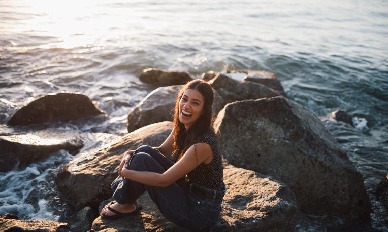 Mandelena Chetty sitting beside a shore on a rock smiling at the camera. 