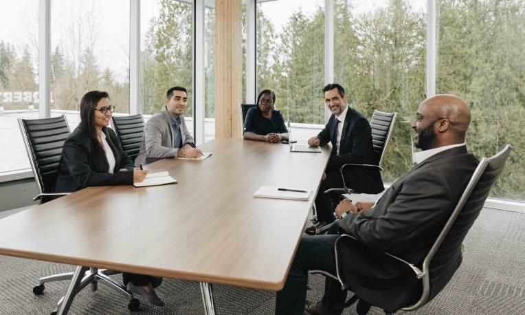 Five adult learners meet around a conference table in a bright, windowed room.