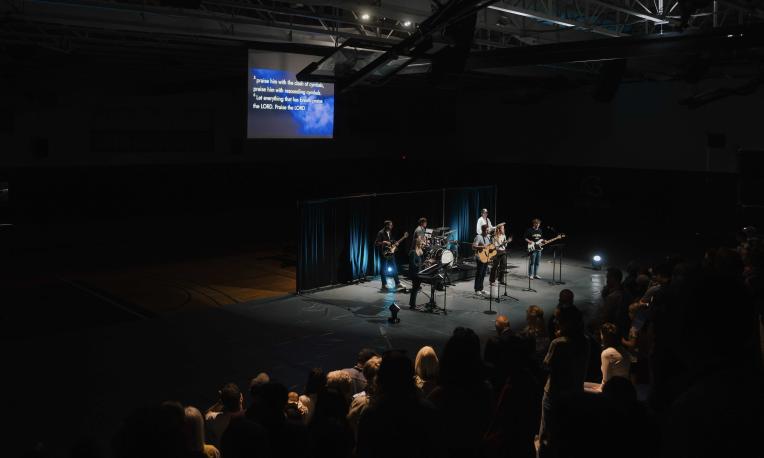 A worship band performs on stage in a dimly lit gym while a crowd gathers during TWU Homecoming.