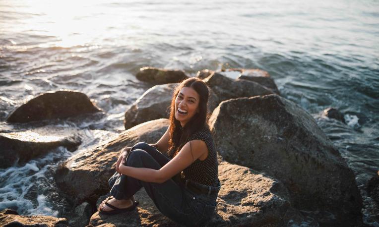 Mandelena Chetty sitting beside a shore on a rock smiling at the camera. 