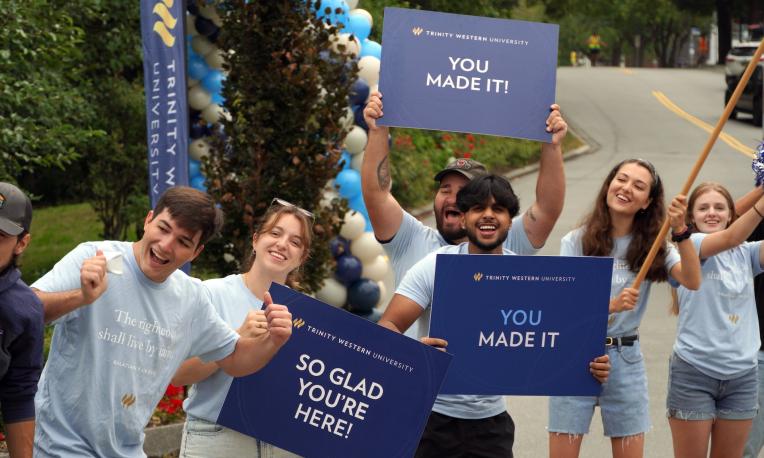 Students and orientation leaders stand along a campus roadway during a TWU orientation event, holding welcome signs and celebrating the arrival of new students on the Langley campus.