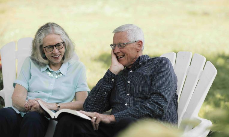 Darwin and Darlis Anderson laughing together while seated outdoors with an open book.
