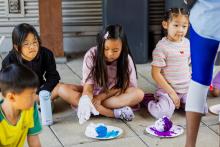 three children sitting on a tiled floor with gloves on doing some painting