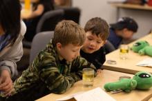 Two young boys hovering over a cup of clear liquid 