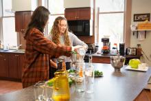 Two students at the Laurentian Leadership Centre are in a kitchen with wooden cabinets and various appliances. One student pours liquid into a bowl held by the other. The countertop features bottles, leafy greens, and a green apple.